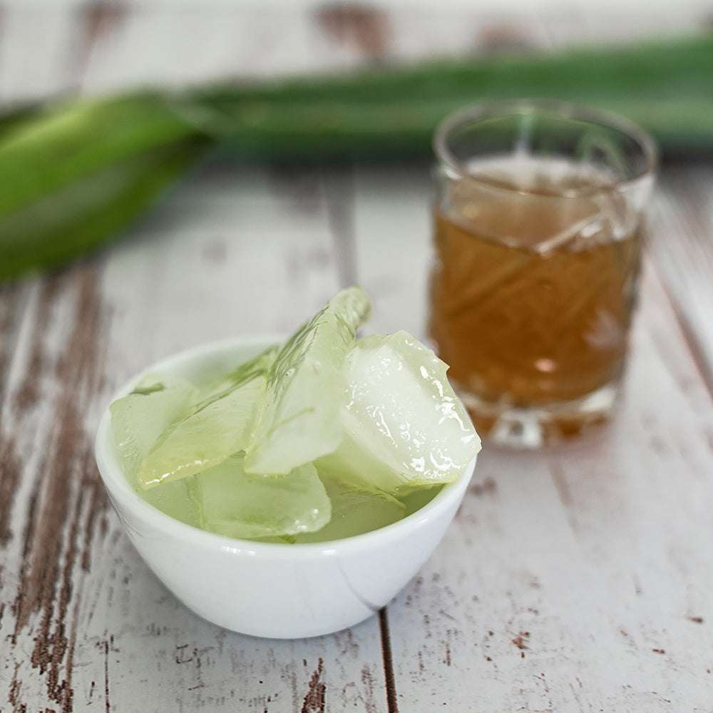 Schale mit klarem Aloe-Vera-Gel mit grünem Farbton, Holzoberfläche, unscharfer Hintergrund mit Aloe-Blatt, Glas mit brauner Flüssigkeit in der Nähe.
