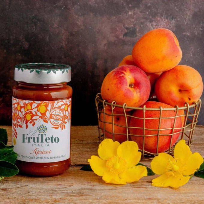 Ein Glas mit der Aufschrift „Frutteto Italia Apricot“, ein mit Aprikosen gefüllter Drahtkorb und daneben leuchtend gelbe Blumen, die auf Bio-Aprikosenaufstrich hindeuten.
