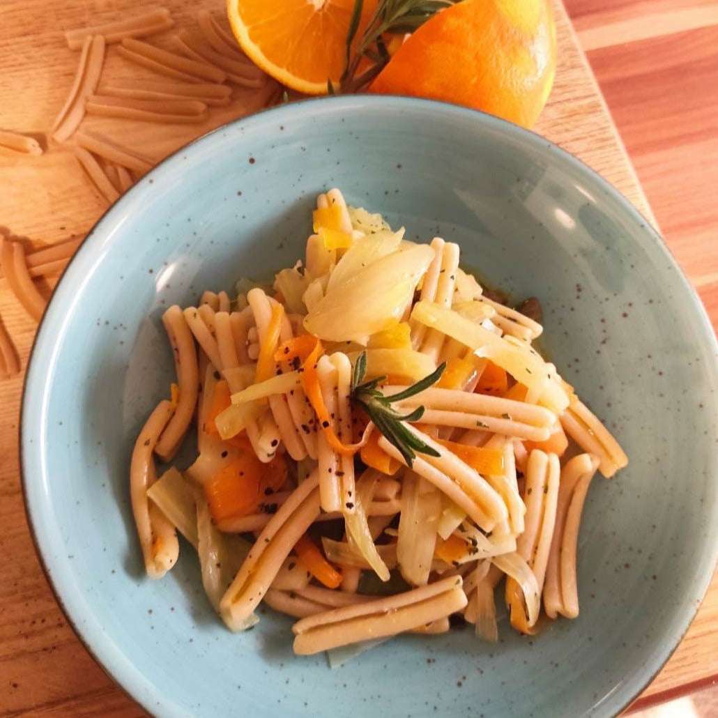 Pasta aus Urgetreide, Caserecce in blauem Teller, garniert mit Orangenscheiben und Rosmarinzweig, auf Holzbrett mit roher Pasta und Orangen.