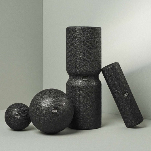 Zwei unterschiedlich große Faszienbälle und zwei Faszienrollen in schwarz, ideal für die Faszienrollmassage, auf grauem Hintergrund.
