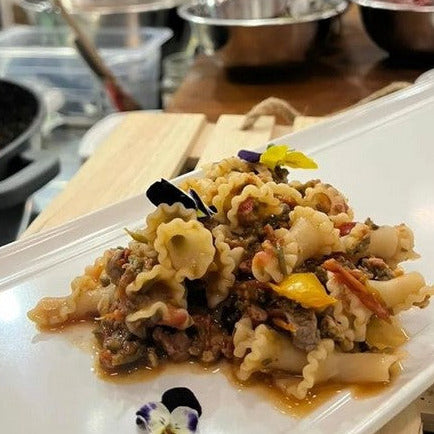 Gigli-Pasta aus Urgetreide, dekoriert mit essbaren Blüten, buntem Gemüse auf weißem Teller, Hintergrund aus Holzbrett.