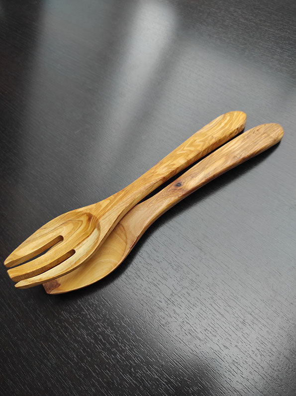 Zwei Salatbesteckstücke aus Olivenholz, elegant gestaltet, liegen auf einer dunklen Holzoberfläche, betonen natürliche Maserung.
