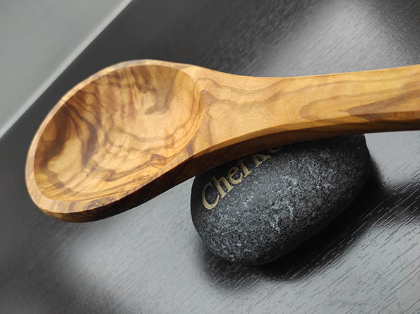 Schöpfkelle aus Olivenholz mit glatter Oberfläche, deutliche Holzmaserung, liegt auf dunkelgrauem Stein auf schwarzem Hintergrund.