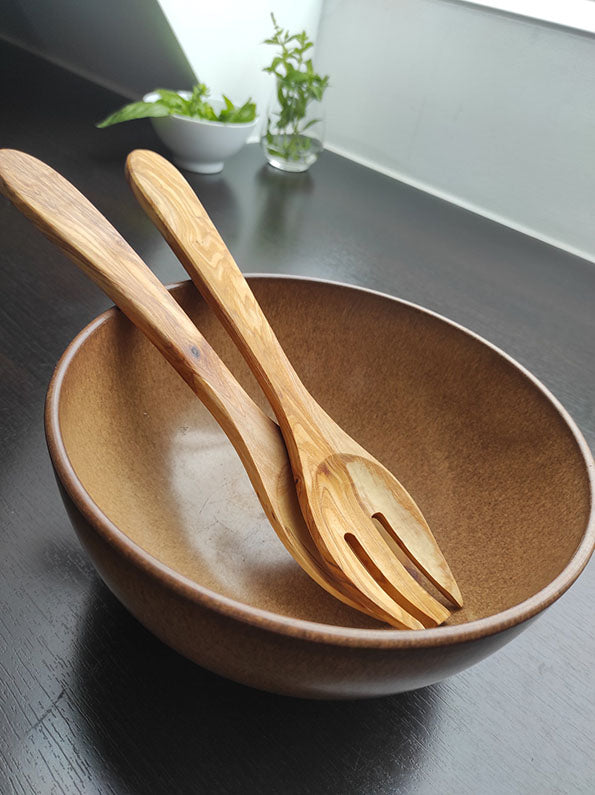 Küchenutensilien Set mit Holzsalatschüssel, passendem Holzlöffel und Gabel auf dunklem Tisch, nahe sonnigem Fenster, Pflanze im Hintergrund.