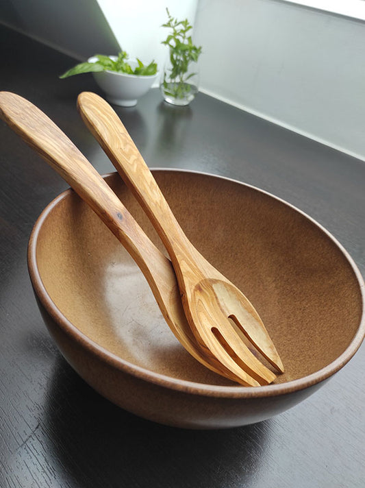 Küchenutensilien Set mit Holzsalatschüssel, passendem Holzlöffel und Gabel auf dunklem Tisch, nahe sonnigem Fenster, Pflanze im Hintergrund.