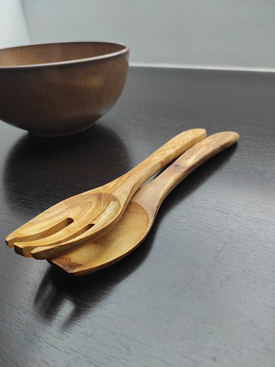 Salatbesteck aus Olivenholz mit Gabel und Löffel, ausgestellt auf einem dunklen Holztisch neben einer braunen Schale.