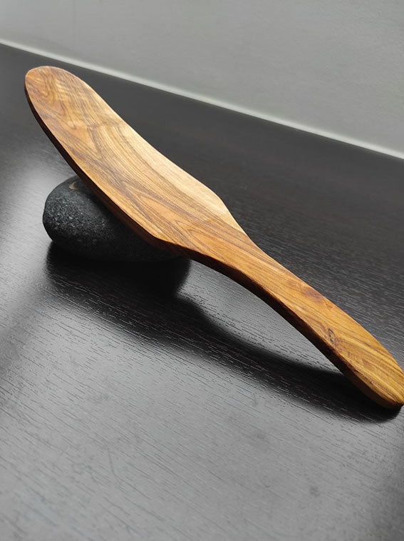 Ovaler Pfannenwender aus Olivenholz, auf einem dunklen Tisch platziert, mit natürlicher Holzmaserung, lehnt auf einem Stein.