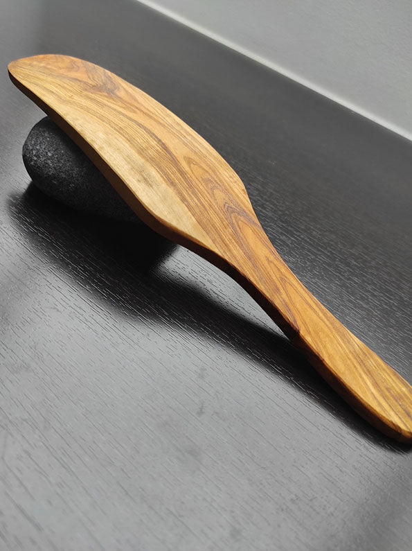 Ovaler Holzspatel aus Olivenholz mit glatter, natürlicher Maserung auf einer dunklen, glänzenden Oberfläche.