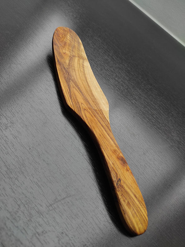 Ovaler Spatel aus Olivenholz auf einer dunklen Holzoberfläche mit glatter, natürlicher Maserung und ergonomischem Griffdesign.
