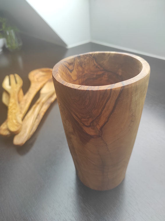 Gefäß aus Olivenholz auf dunkler Oberfläche, zeigt lebhafte Maserungen, mit passendem Olivenholz-Besteck im Hintergrund.