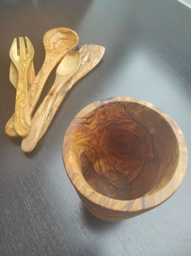 Set aus Küchenutensilien aus Olivenholz, umfasst Gabel, Löffel und einen Becher, präsentiert auf dunklem Holzuntergrund.