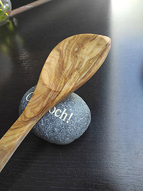 Olivenholz-Kochlöffel mit markanter Maserung auf einem gravierten Stein liegend, dunkler Hintergrund, naturbelassenes Holz.