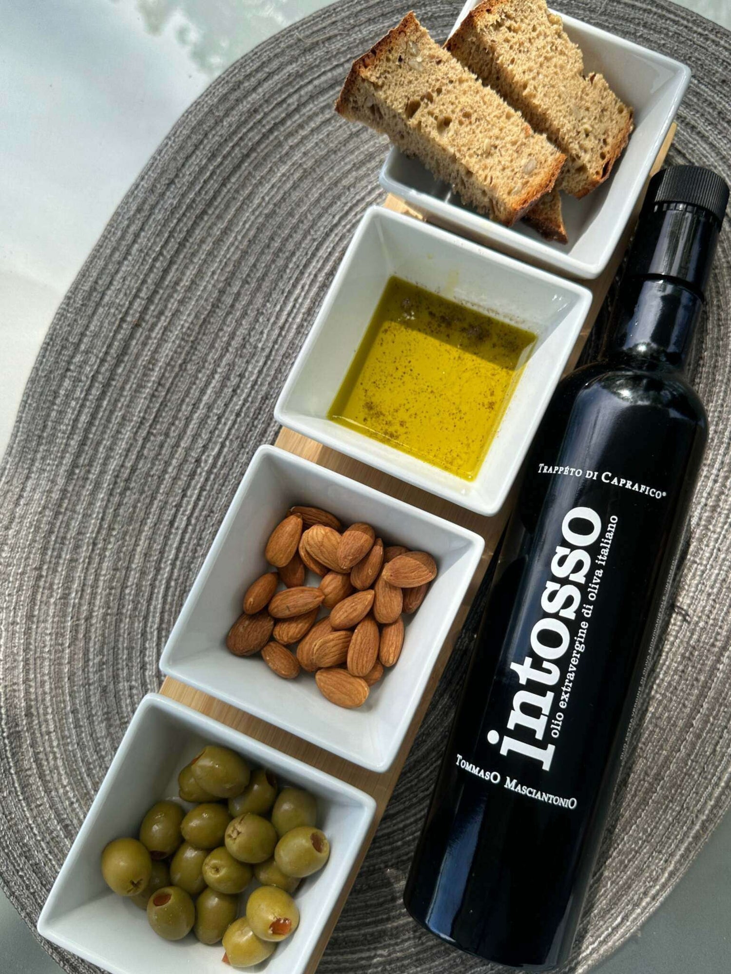 Feinkost Geschenkset mit Olivenölflasche, Brotscheiben, aromatisiertem Dip-Öl, Mandeln und grünen Oliven auf elegantem Tischset.