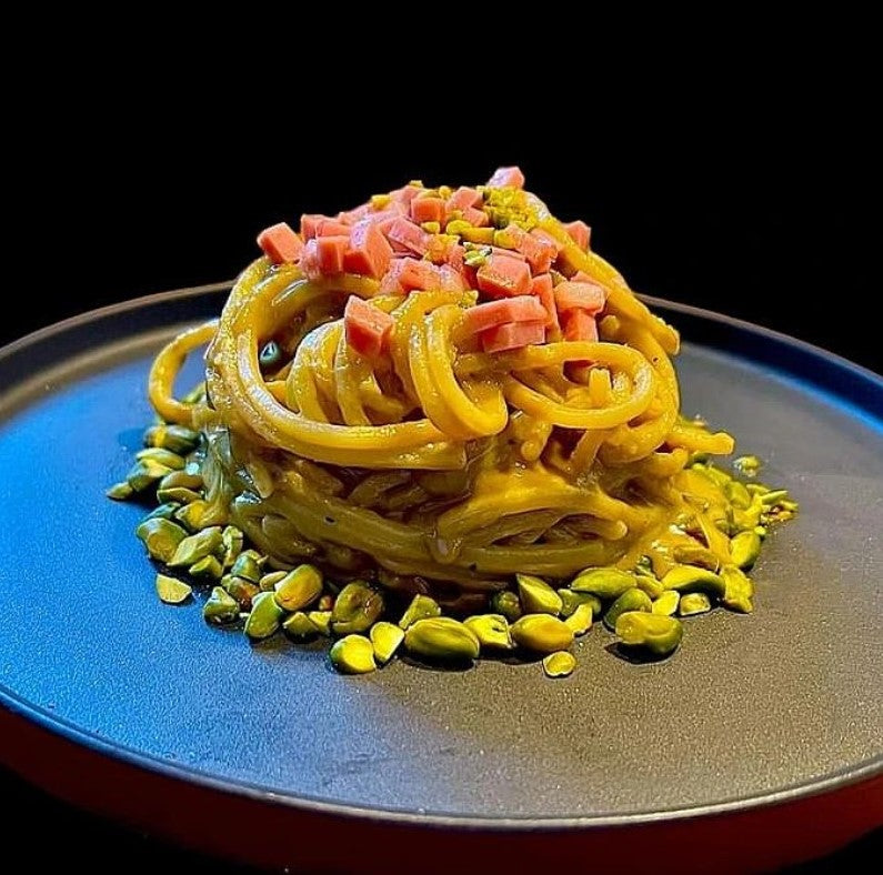 Spaghetti quadrati aus Urgetreide, serviert mit Würfeln von Schinken und gehackten Pistazien auf einem schwarzen Teller.