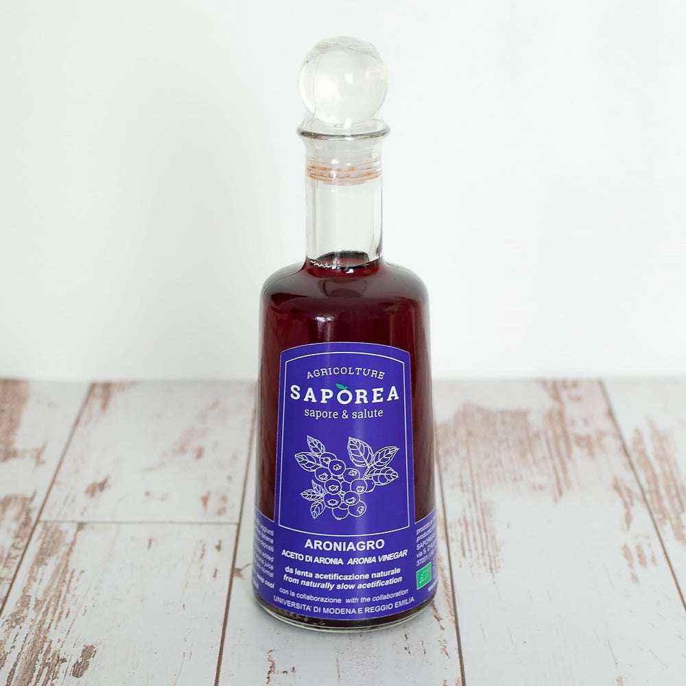 Glasflasche Aronia-Essig mit rundem Verschluss, blauem Etikett mit botanischen Illustrationen, auf rustikalem Holzuntergrund.