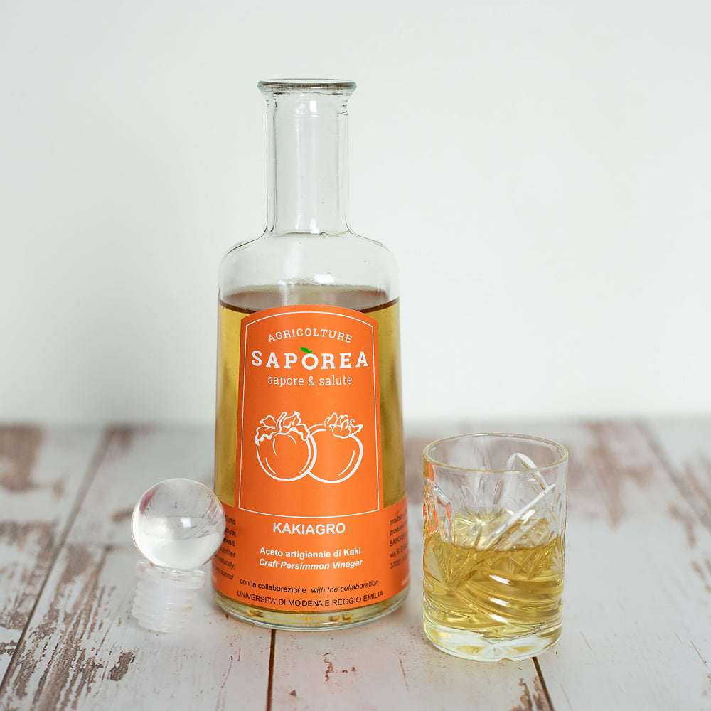 Eine klare Flasche Kaki-Essig mit orangefarbenem Etikett mit Kaki-Motiven und einem Glas, das mit dem Essig gefüllt ist.