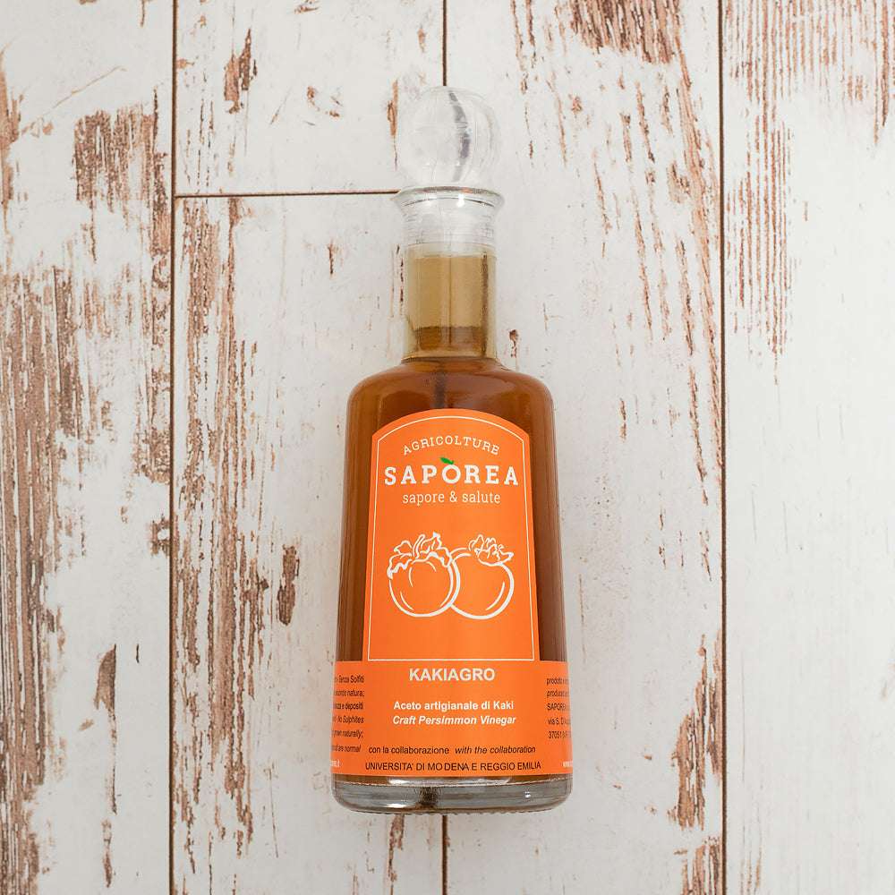 Flasche Kaki-Essig mit orangefarbenem Etikett und stilisierten Kakis, auf rustikalem Holzboden platziert, zeigt elegantes Design.