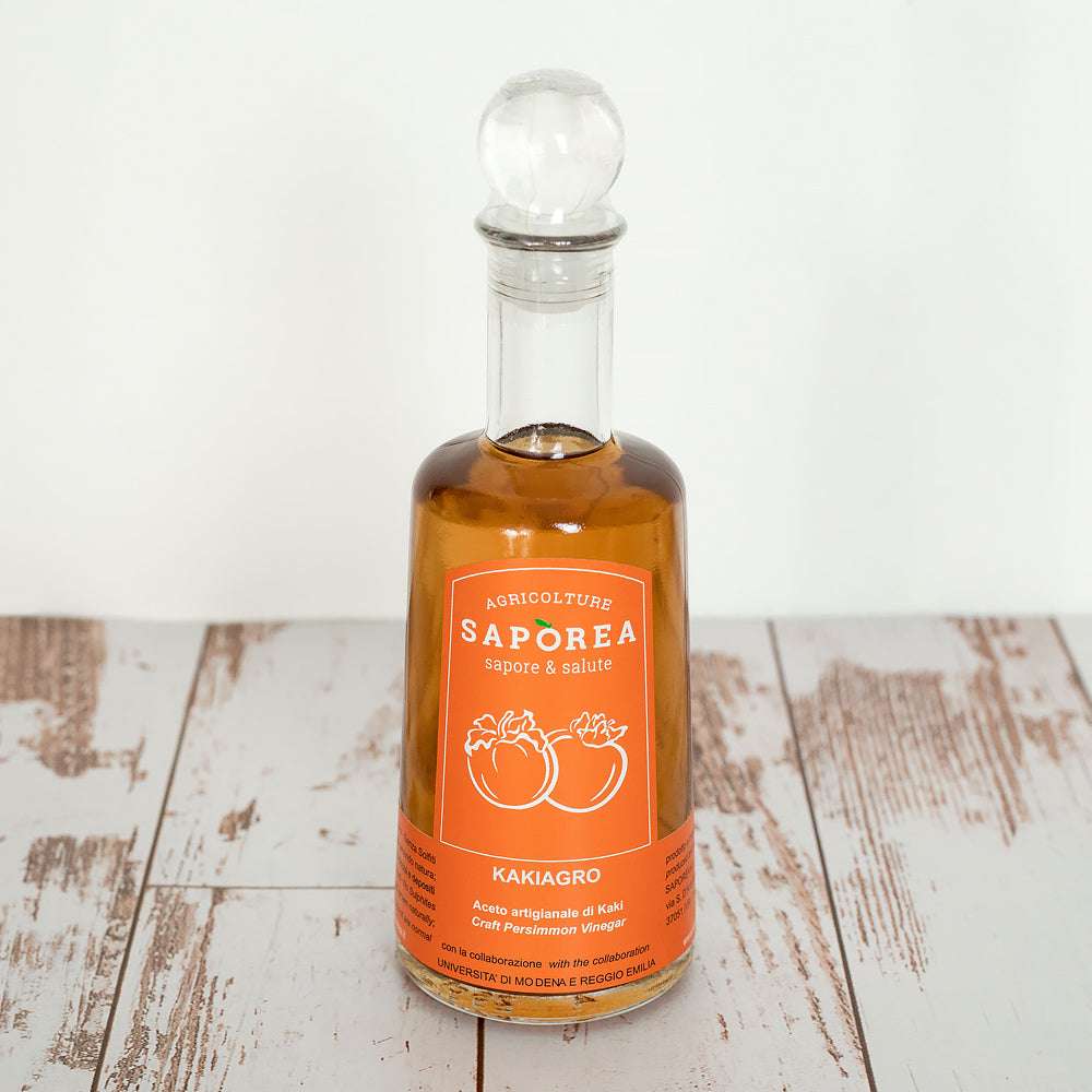 Eine klare Glasflasche Kaki-Essig mit orangem Etikett, darauf zwei Kaki-Früchte, auf rustikalem Holztisch platziert.