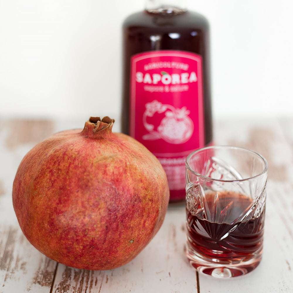 Granatapfel-Essig-Flasche mit leuchtend rotem Etikett, daneben ein frischer Granatapfel, halbgefülltes Kristallglas. Rustikale Holzoberfläche.