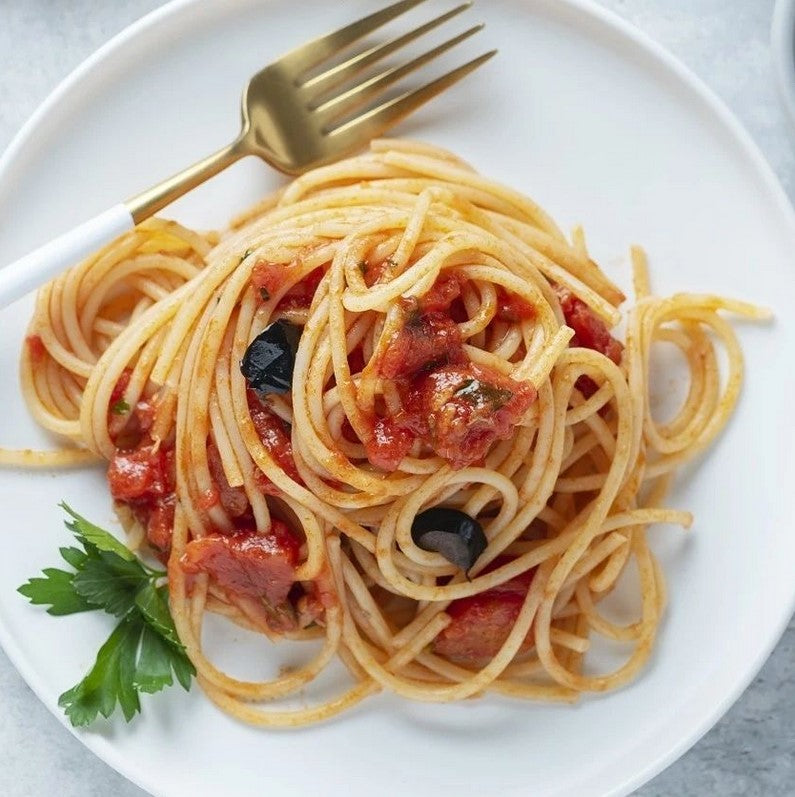 Spaghetti aus Urgetreide auf weißem Teller, serviert mit Tomatensauce, schwarzen Olivenstücken und Petersiliengarnitur.