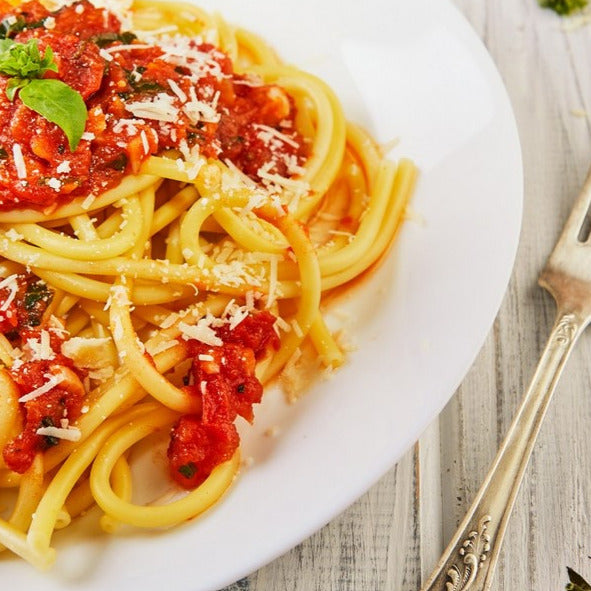 Ein Teller mit italienischer Pasta in Tomatensoße, garniert mit frischem Basilikum und geriebenem Käse, neben einer Gabel.