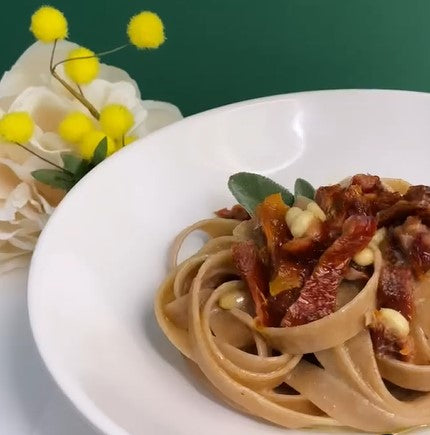 Tagliatelle aus Urgetreide auf einem Teller, garniert mit getrockneten Tomaten, Kräutern und einer Blume an der Seite.