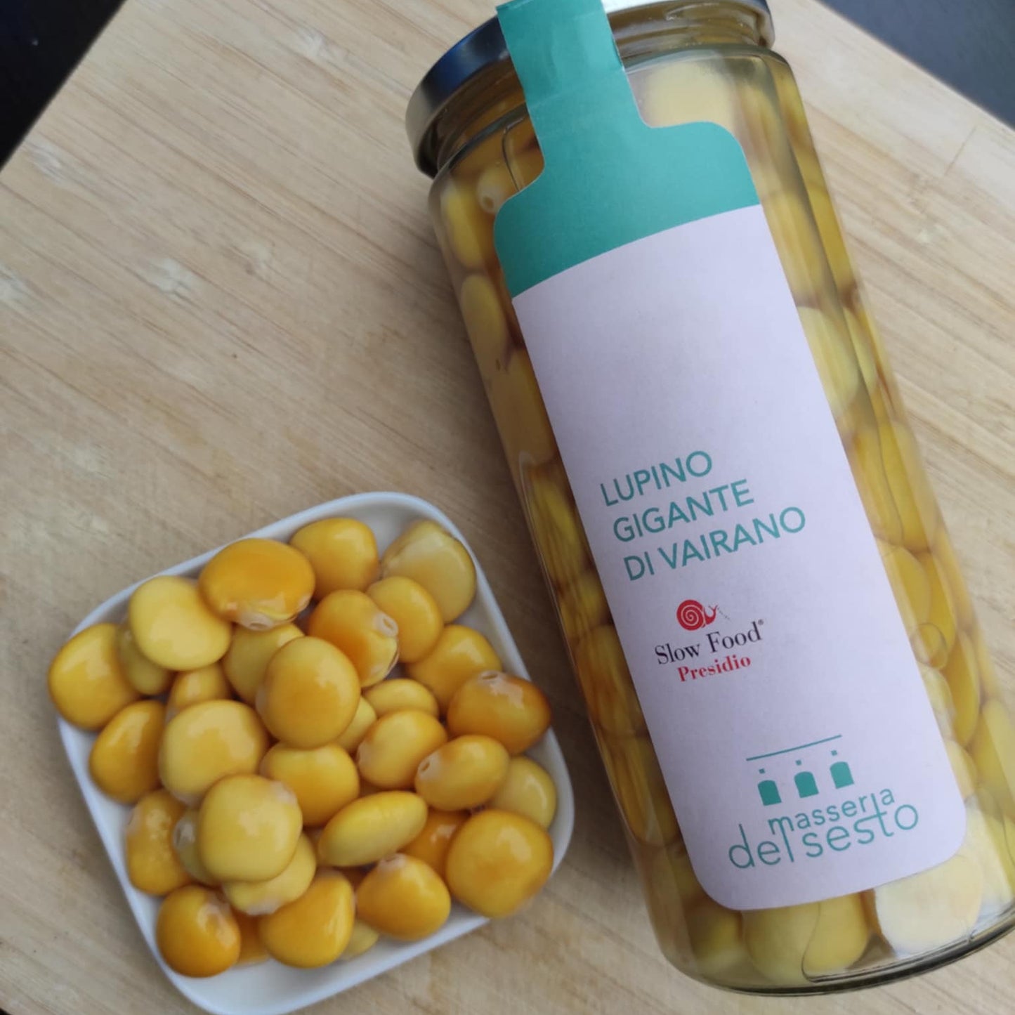 Ein Glas mit der Aufschrift "Lupino Gigante di Vairano" gefüllt mit gelben Lupinen, daneben eine weiße Schale mit Lupinen.