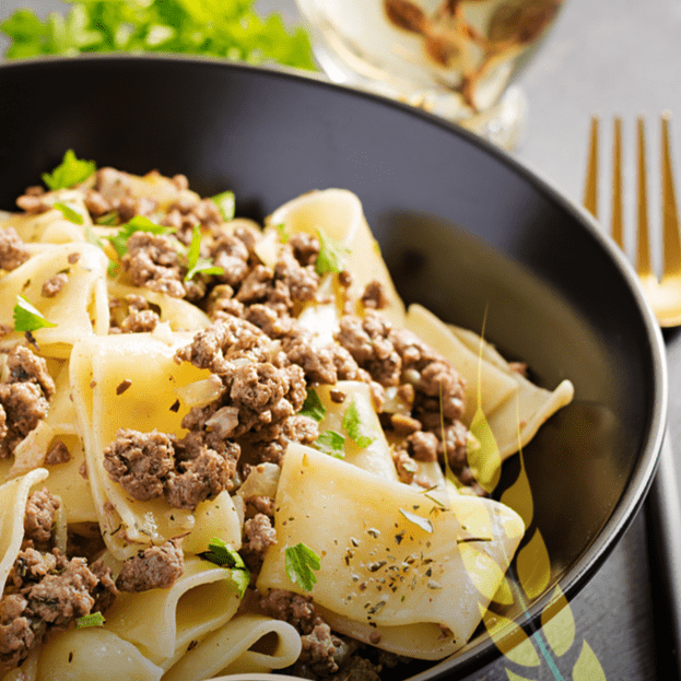 Schwarze Schale mit calamari-förmigen Nudeln aus alten Getreidesorten, belegt mit Hackfleisch und frischen Kräutern, neben einer Gabel platziert.