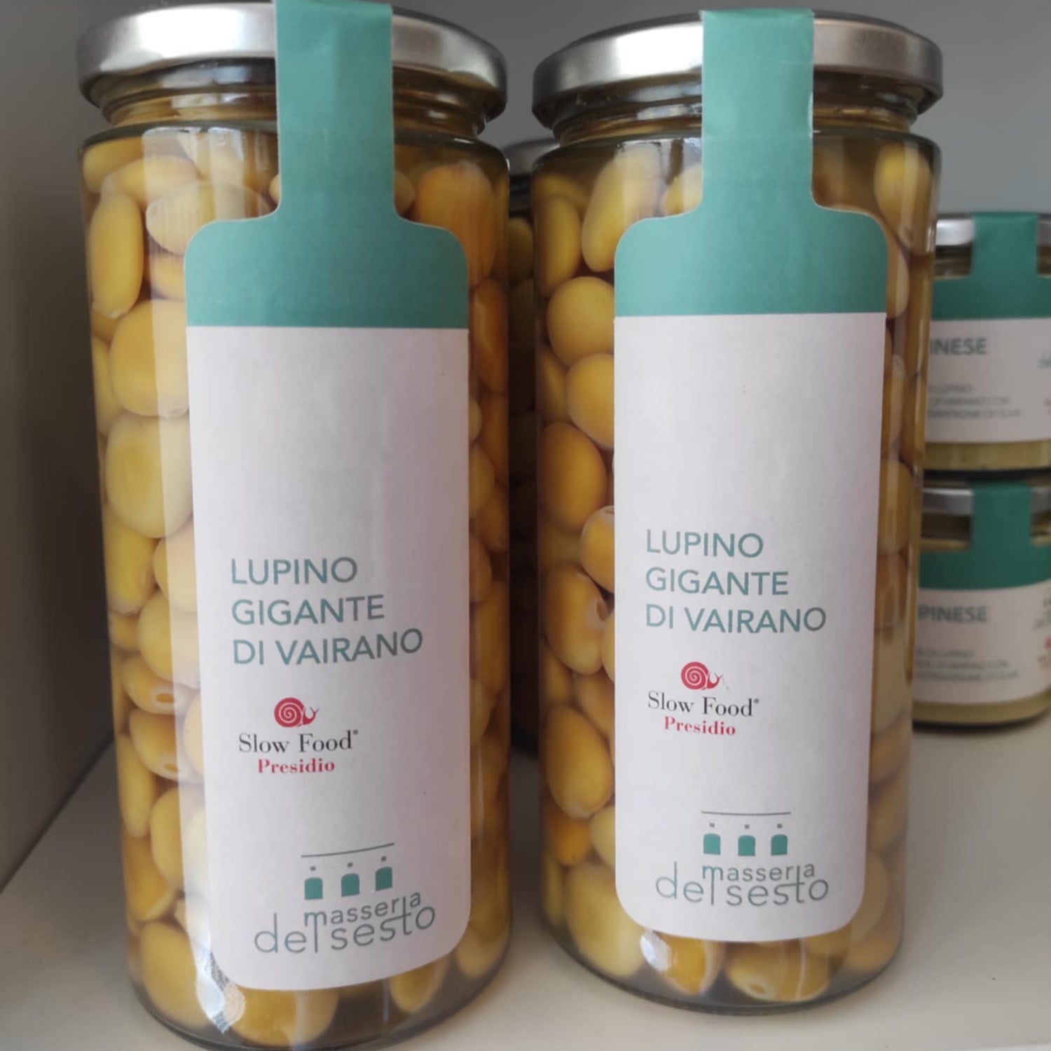 Zwei Gläser mit gelben Lupinen aus Vairano in Salzlake, beschriftet mit "Lupino Gigante di Vairano", auf einem Regal.