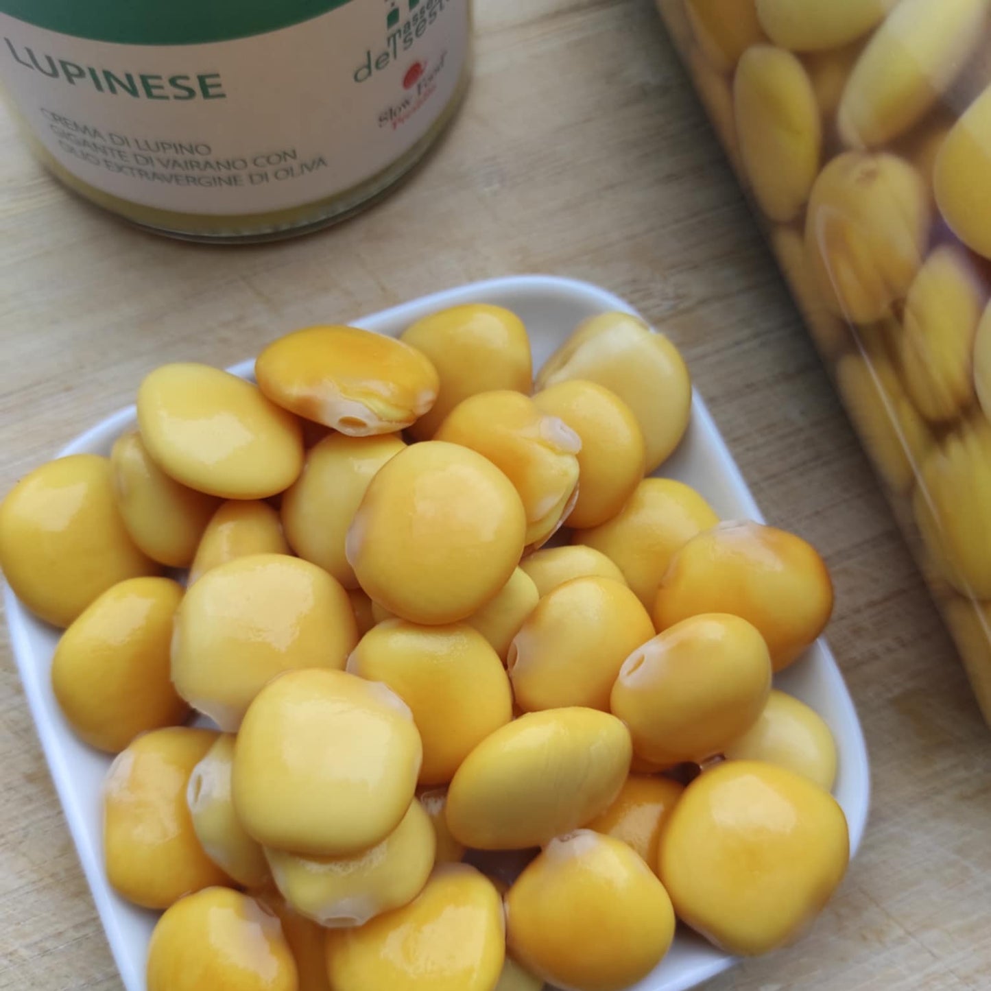 Eine kleine weiße Schale, gefüllt mit leuchtend gelben Lupinen in Salzlake; im Hintergrund sind ein Glas und eine Dose Lupinen aus Vairano zu sehen.