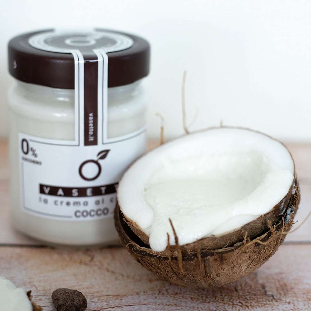 Ein Glas Kokoscreme "Kokos 0% Zucker*" mit braunem Deckel neben einer halbierten Kokosnuss auf heller Holzoberfläche.