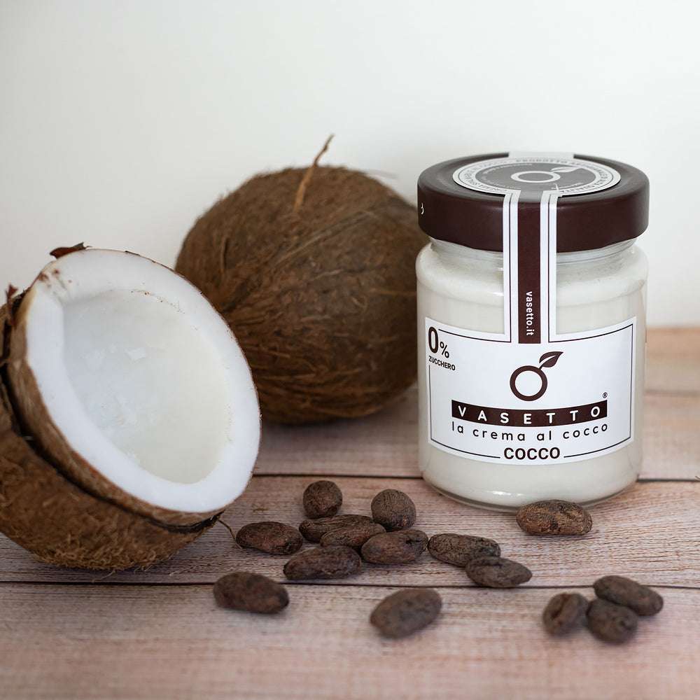 Ein Glas "Kokos 0% Zucker*" auf einer Holzoberfläche, umgeben von halbierten Kokosnüssen und verstreuten Kakaobohnen.