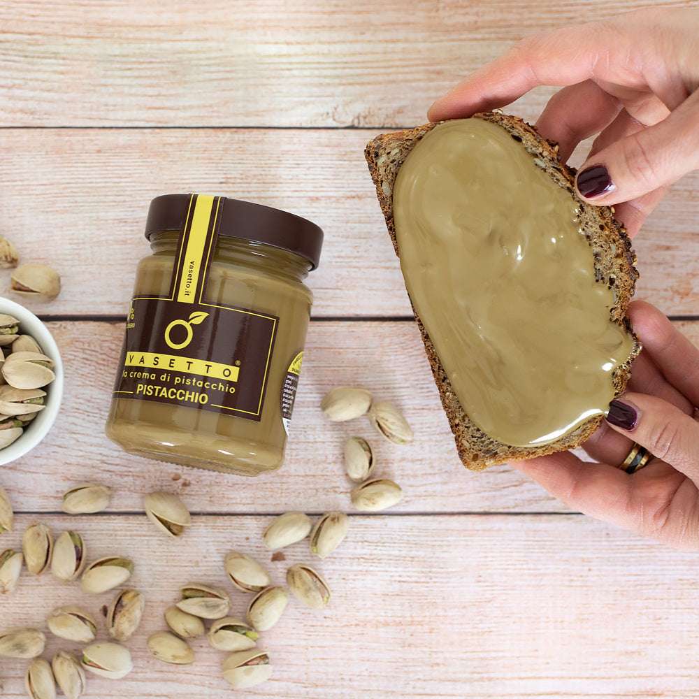 Ein Glas Pistaziencreme mit der Aufschrift "Pistachio", verstreute Pistazien, eine Hand hält Brot bestrichen mit der Pistaziencreme.