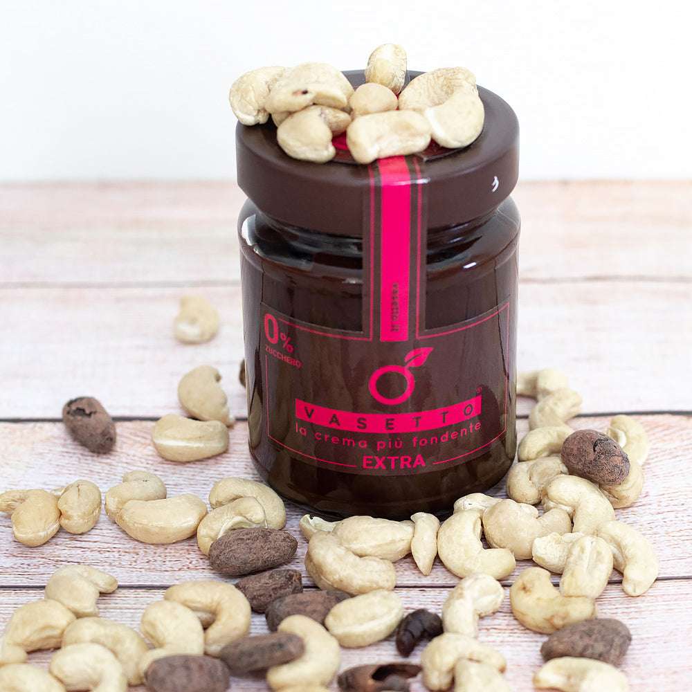 Ein Glas mit dem Brotaufstrich „Cashew 0% Zucker*“, mit dunklem Etikett, umgeben von verstreuten Cashewnüssen auf einer Holzfläche.