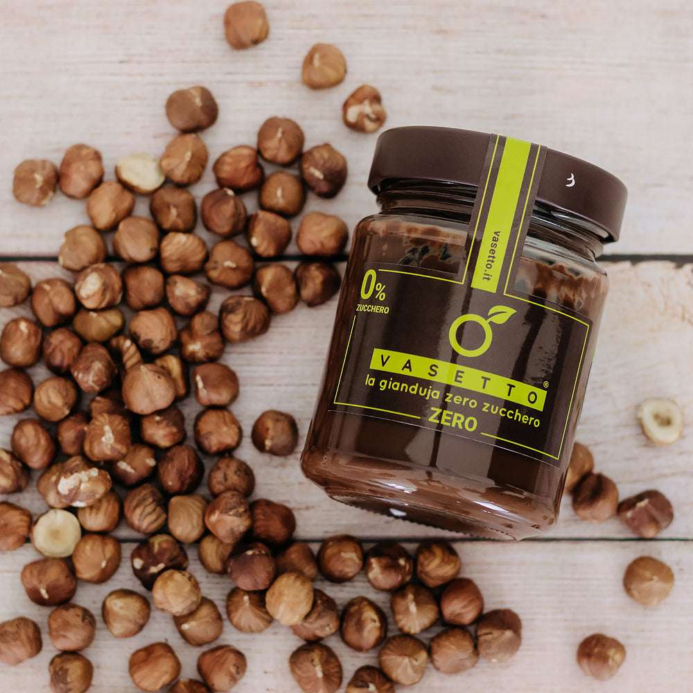 "Vasetto" zuckerfreier Gianduja-Schokoladenaufstrich, umgeben von verstreuten Haselnüssen auf einer Holzoberfläche.