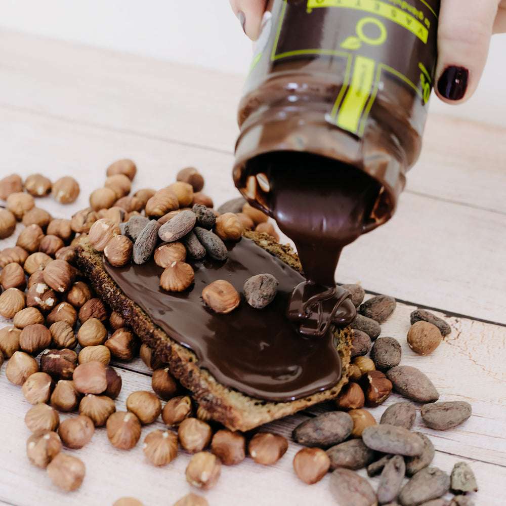 Ein Glas Haselnuss 0% Zucker*" Schokoladenaufstrich wird auf eine Scheibe Brot gegossen, umgeben von Haselnüssen und Kakaonibs.