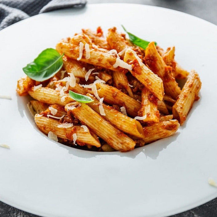 Ein Teller mit Vollkorn-Penne aus Urgetreide, in reichhaltiger Tomatensoße, garniert mit geriebenem Käse und frischen Basilikumblättern.