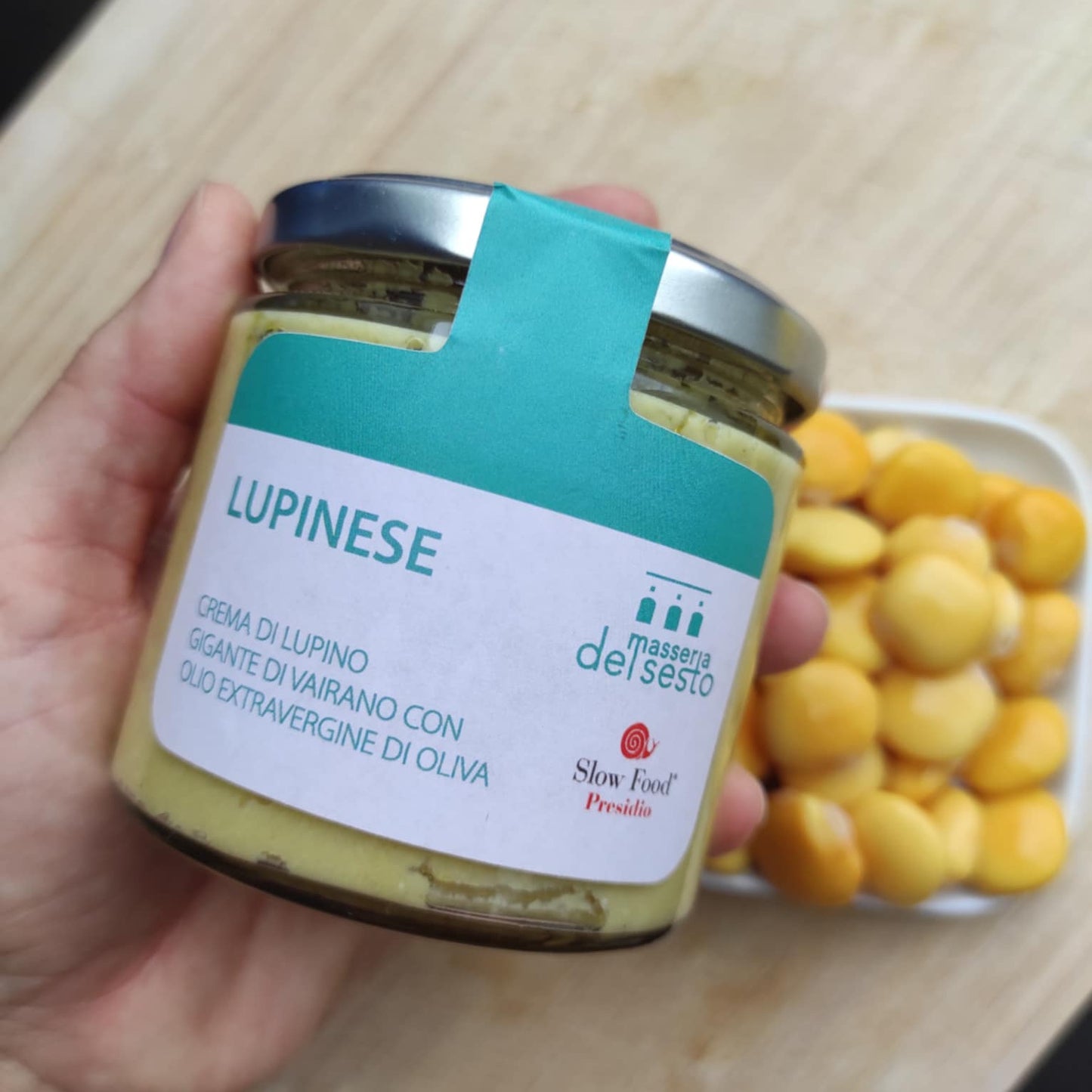 Hand hält Glas Lupinese Creme aus Lupinen, 180g, auf holzartigem Hintergrund, daneben eine Schale mit gelben Lupinenbohnen.